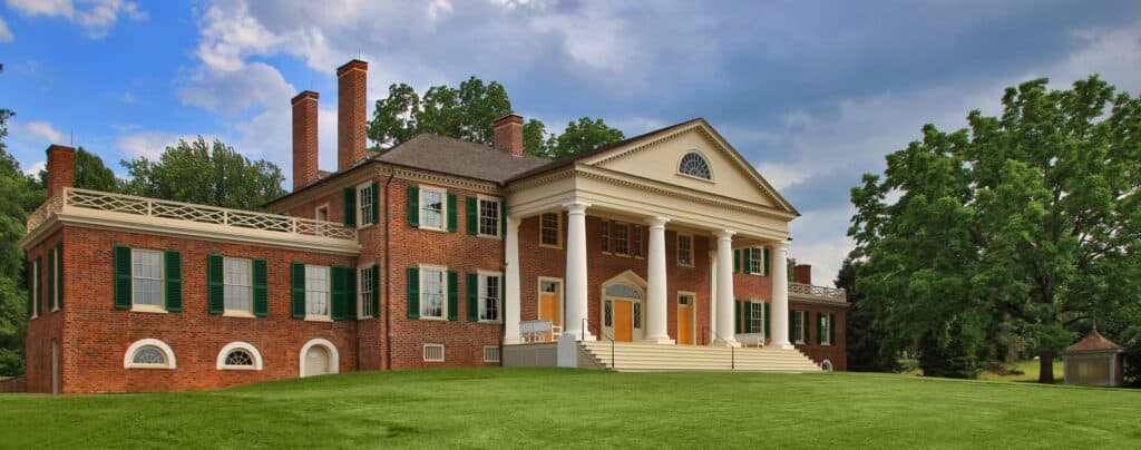 A large, two-story brick mansion with white columns and green shutters stands on a well-manicured lawn under a blue sky with clouds. Trees surround the building.