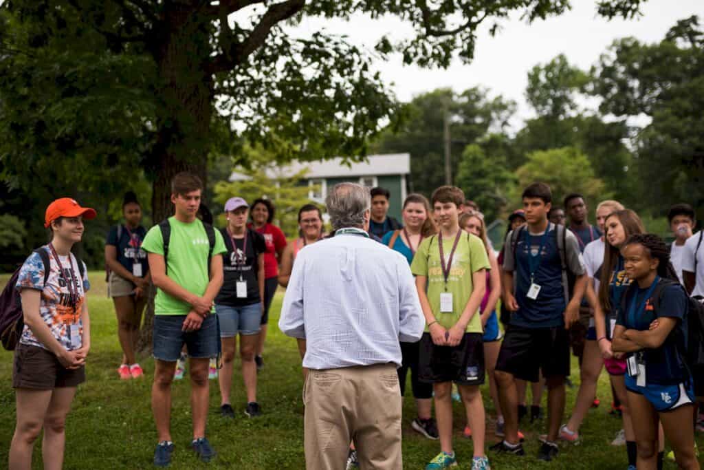 A person stands outdoors facing a group of individuals, who are attentively listening. The group appears to be participating in an outdoor activity or lecture. Trees and grassy area are visible.