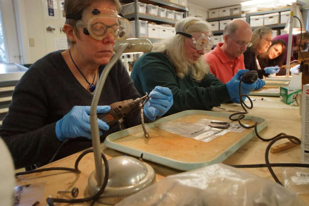 Several people wearing safety goggles and gloves use tools to work on objects at a table in a workshop.