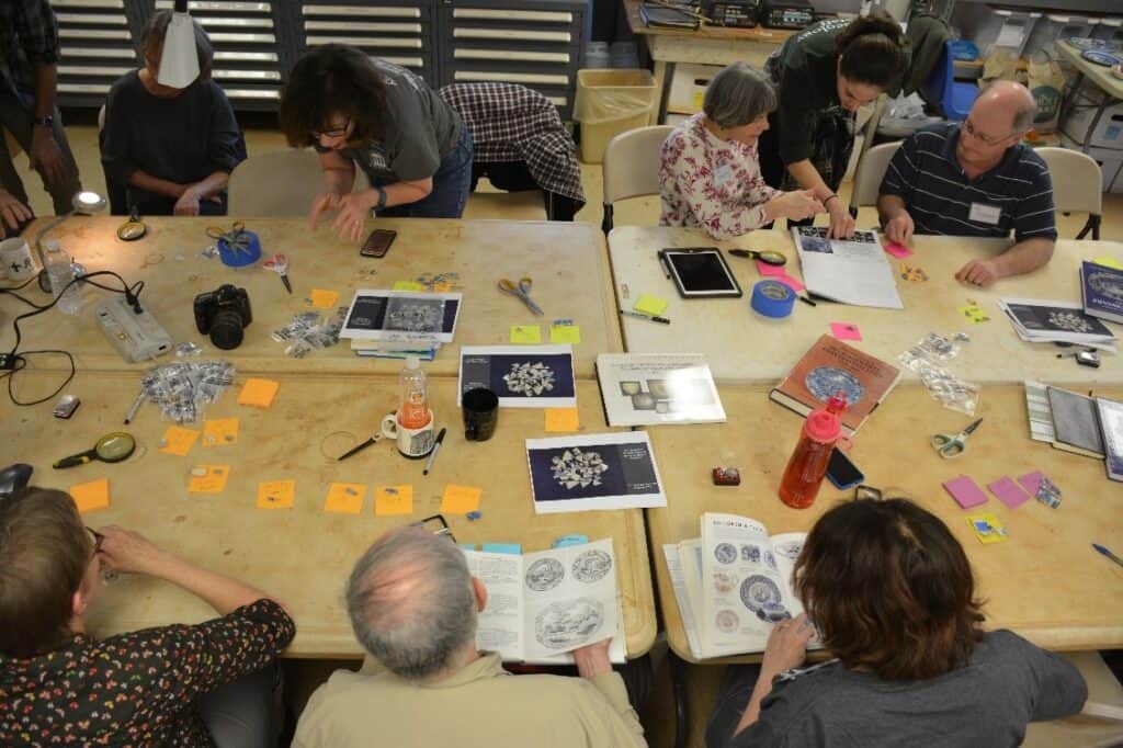 Group of people sitting around a table working on crafts and reading books. Various tools, papers, and small items are scattered across the table.