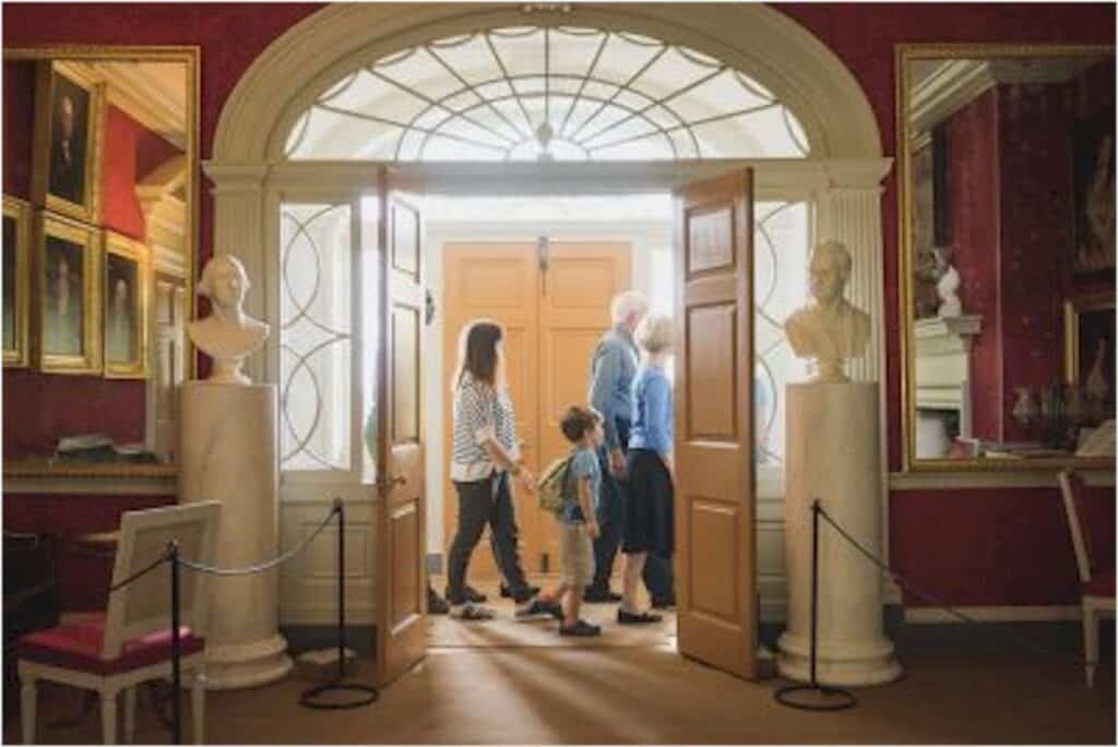 A group of people, including children, walk through a doorway in a historical building adorned with statues and framed portraits.