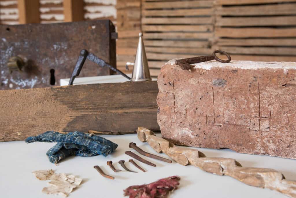 A collection of historical artifacts including an old brick, rusty nails, a twisted wooden piece, scraps of fabric, and a metal tool, displayed on a table with a wooden backdrop.