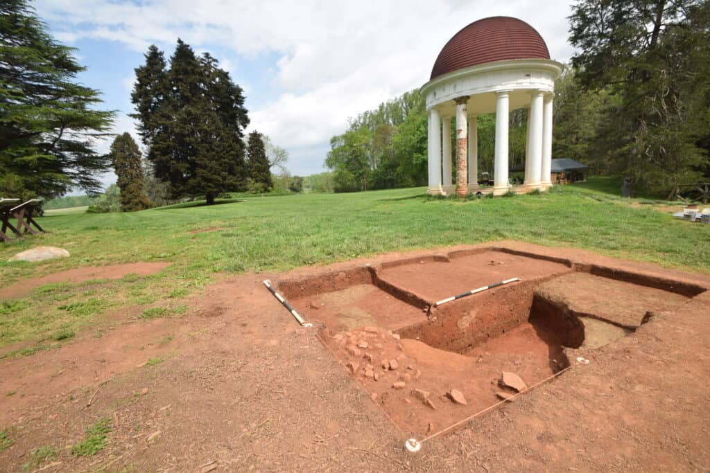 An archaeological excavation site with a partially uncovered structure in foreground; in background, a white circular columned rotunda with a domed roof stands on a grassy area surrounded by trees.
