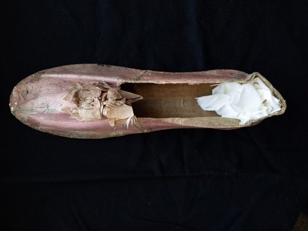 A worn-out pink ballet shoe with visible tears and white tissue paper stuffed inside, displayed on a black background.