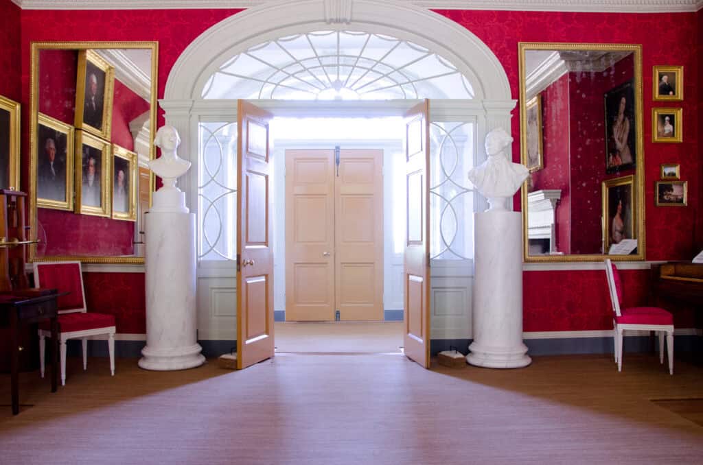 A room with vibrant red walls, adorned with framed portraits and sculptures. Large double doors are open, revealing a bright hallway in the background. Two white busts stand on pillars by the doorway.
