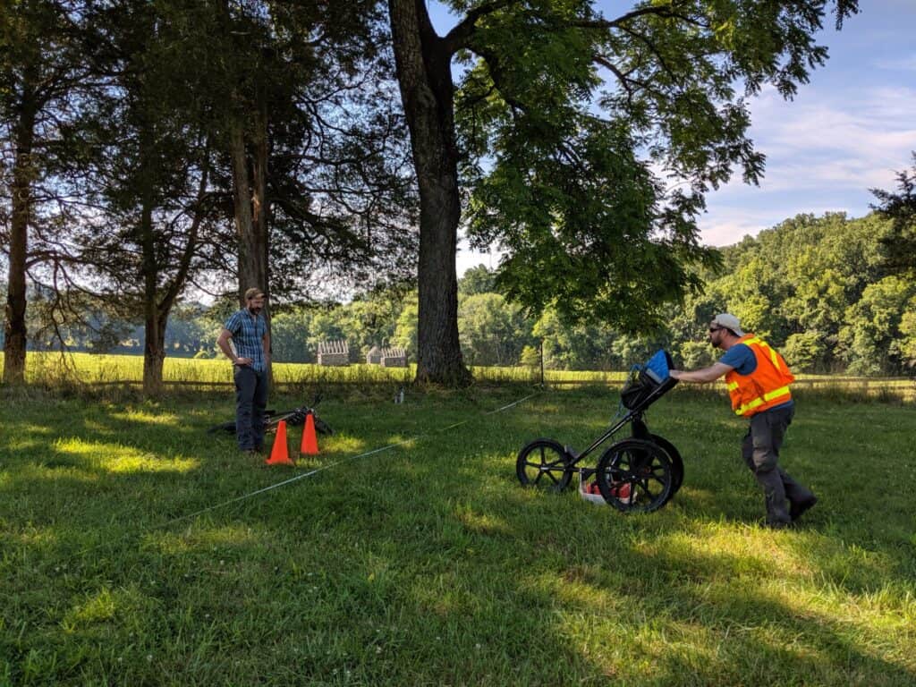 Two men conducting ground-penetrating radar (GPR) survey on a grassy field, one man operates the equipment while another observes. The scene is set outdoors with trees and an open field in the background.
