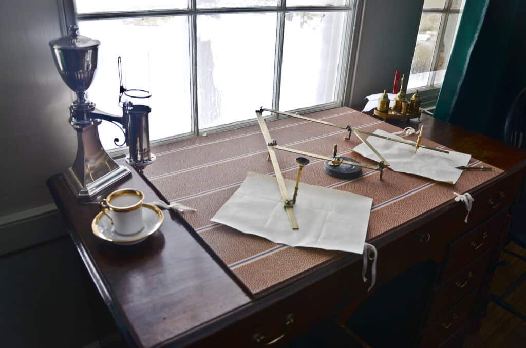 An old-fashioned drafting table with drawing tools, a cup of coffee, and papers, situated next to a window.