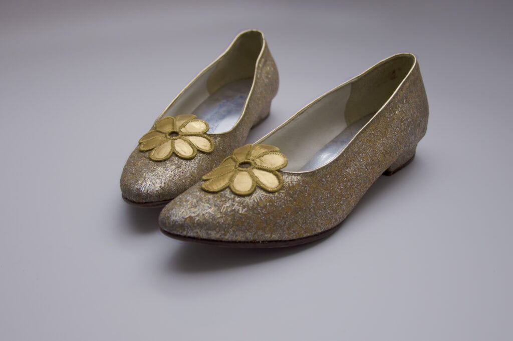 A pair of golden glitter shoes with large yellow flowers on the toes, displayed on a plain grey background.