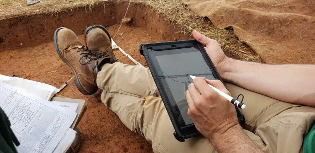 A person sitting on the ground uses a stylus to draw on a tablet. Nearby, there are papers with handwritten notes.
