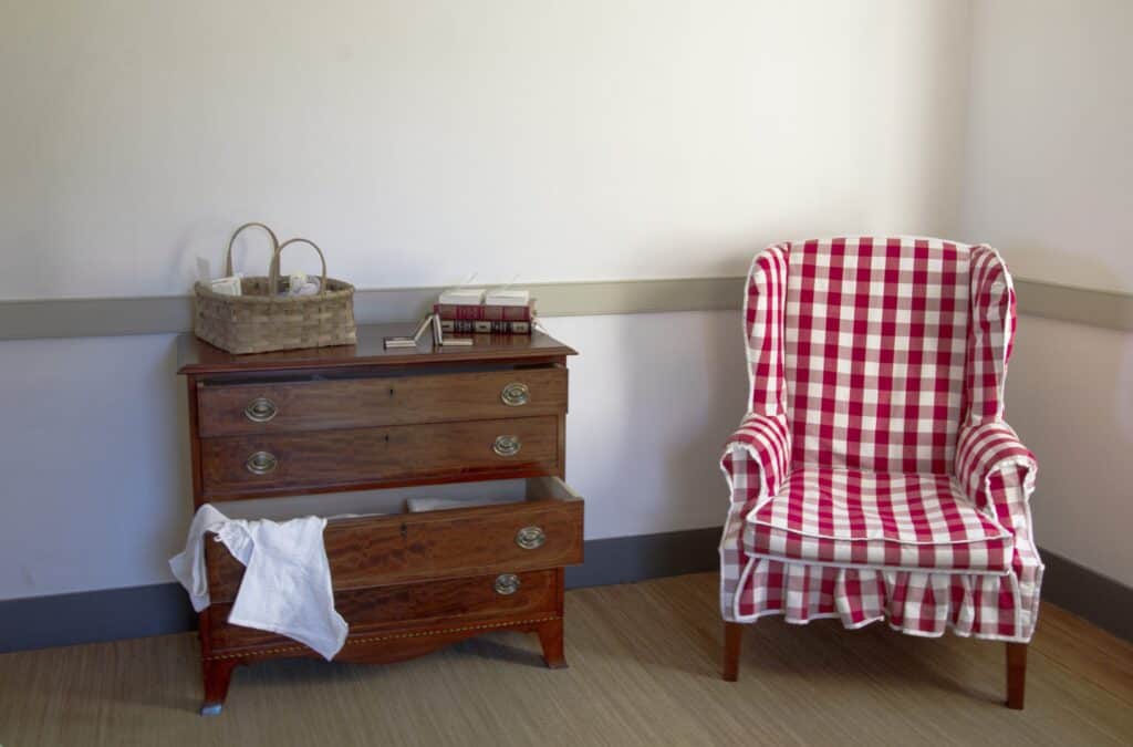 A room with a wood dresser with open drawers, a basket on top, and a red-and-white checkered armchair beside it.