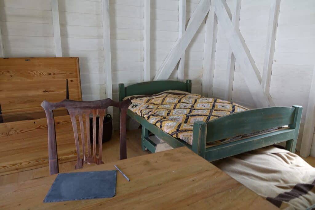 A simple room features a green wooden bed with a quilt, a wooden chair missing its seat, a wooden table with a slate and pencil, and wooden boards propped against the white plank walls.
