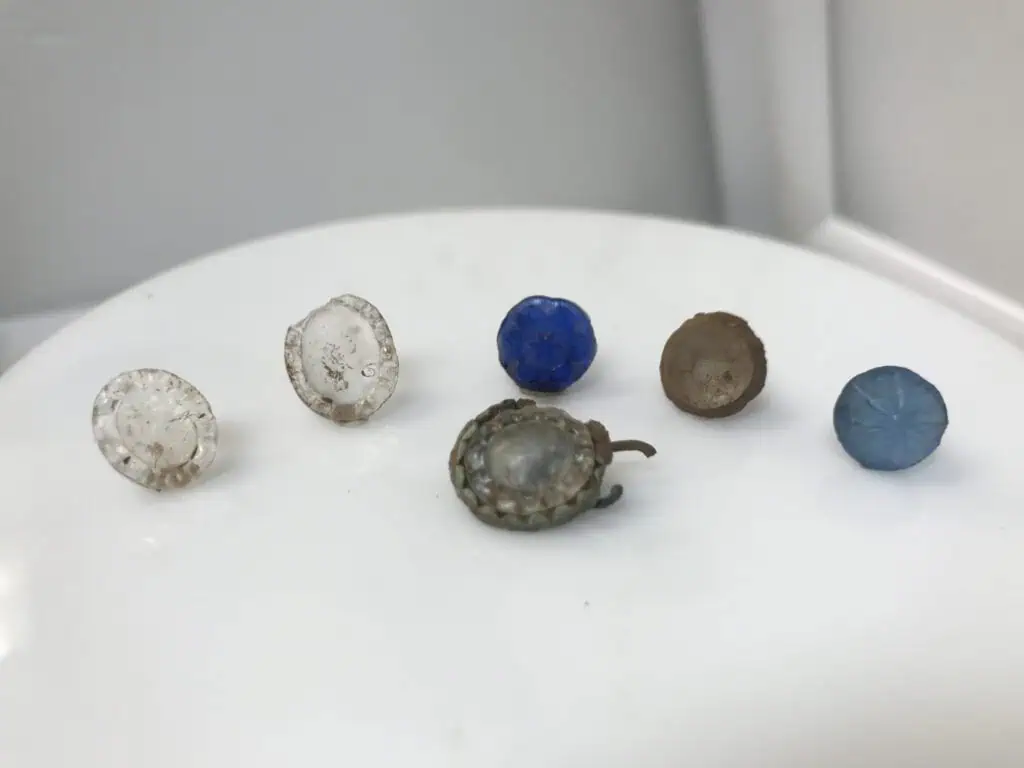 Six small, round buttons of various colors and materials, including clear, blue, and brown, are displayed on a white surface.