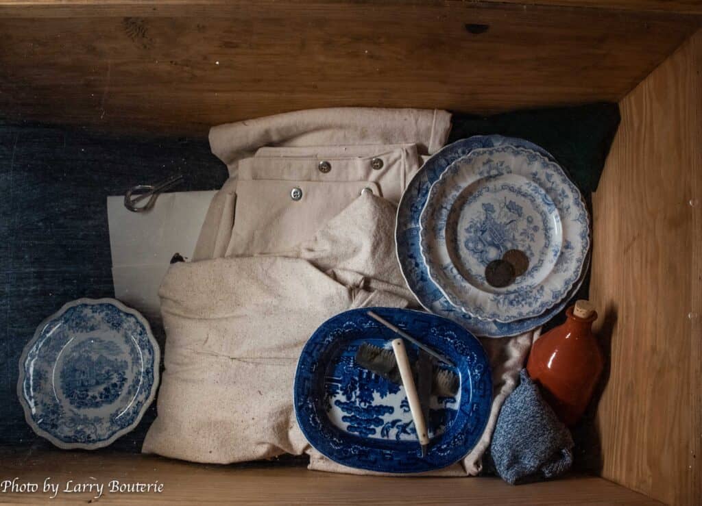 Wooden box containing vintage blue and white dishes, a small brown jug, coins, a grey knitted item, a brush, and light-colored folded pants.