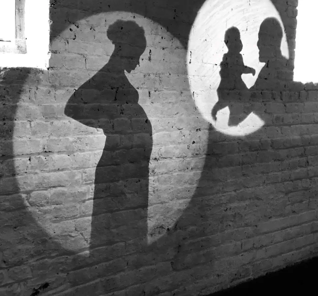 Silhouettes on a brick wall show a pregnant woman in one circle of light and the same woman holding a baby in another, suggesting life stages.