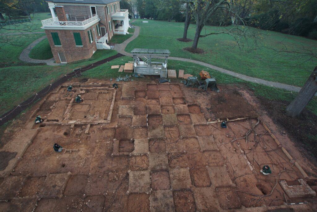 An archaeological excavation site in front of a large brick building, showing a grid of cleared earth and tools scattered around the area.