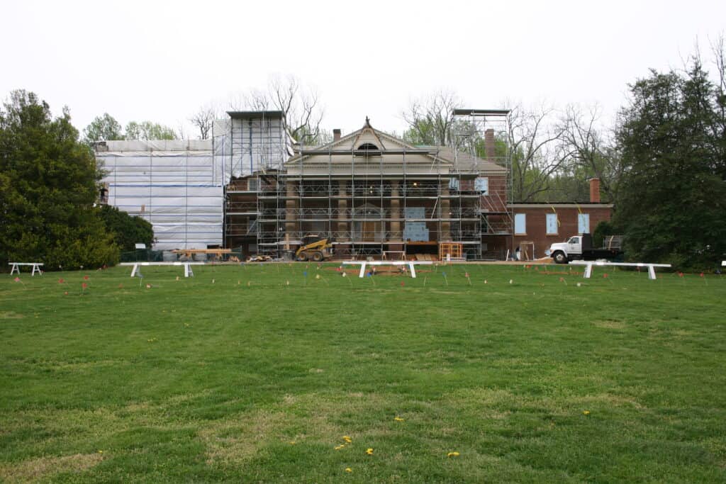 A large house under renovation with scaffolding and construction materials on the front lawn. Trees flank both sides of the building, and a vehicle is parked nearby.