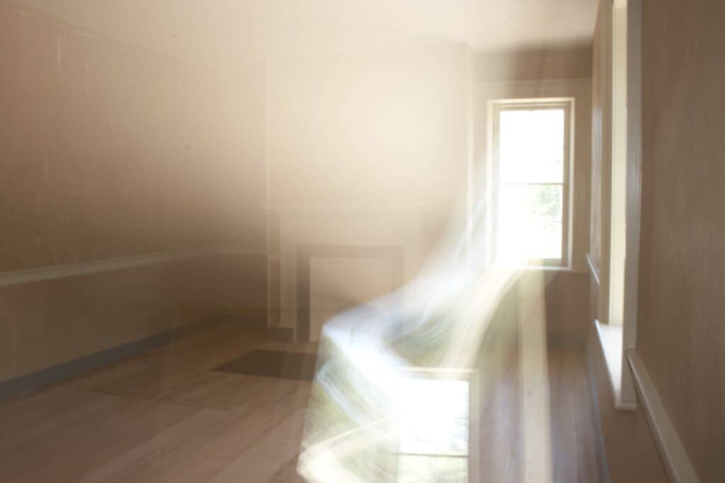 A dimly lit, empty room with a small window letting in light, and a ghostly, translucent figure or light trail near the center.