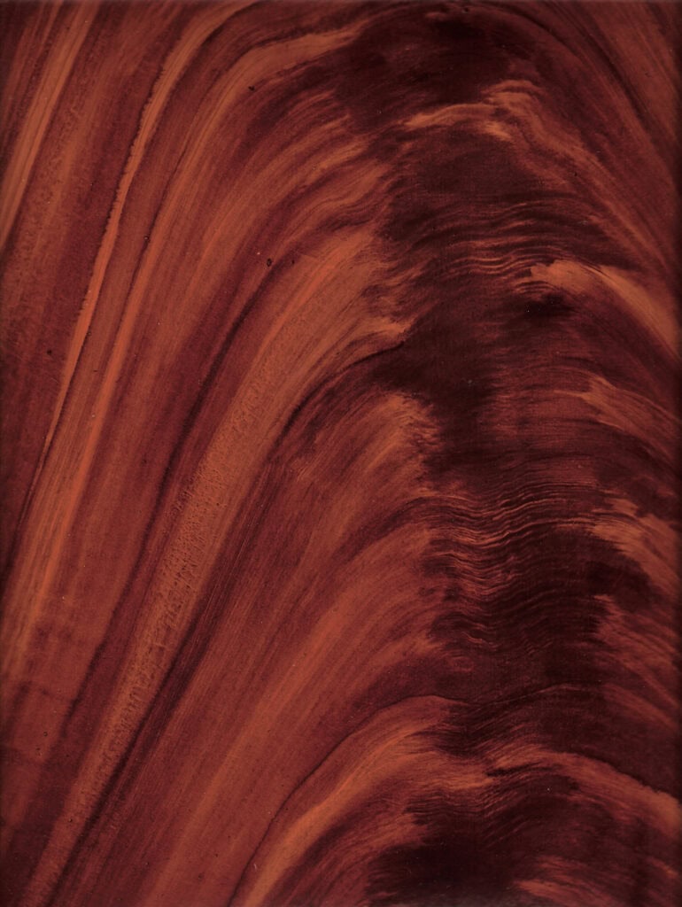 A close-up view of polished, reddish-brown wood grain with natural, wavy patterns.
