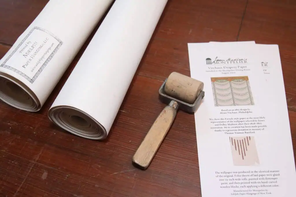 Two rolls of paper, a wooden-handled ink roller, and a printed sheet with decorative designs are displayed on a wooden surface.