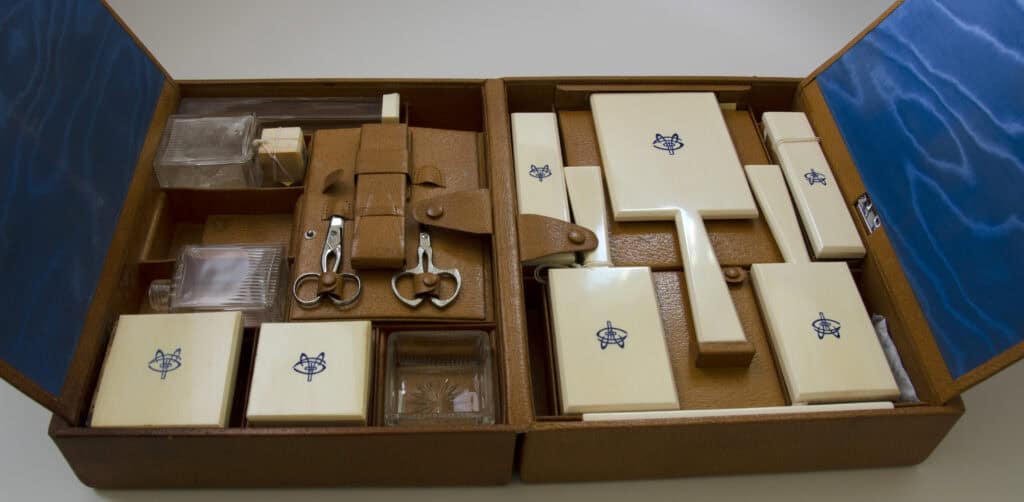 Open beige travel toiletry set in a blue-lined case, includes scissors, combs, nail tools, and various rectangular containers with blue emblems.