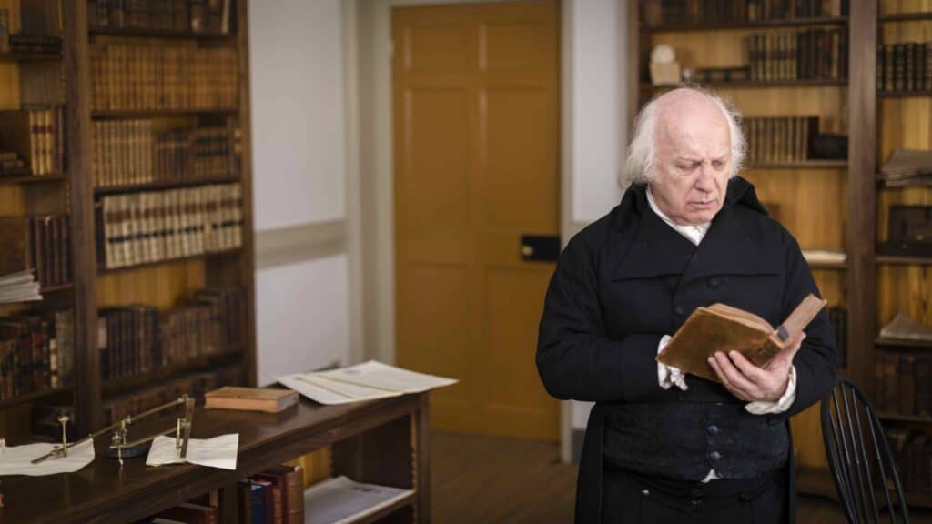 An elderly man with white hair and in period clothing stands in a study with bookshelves, reading a book.