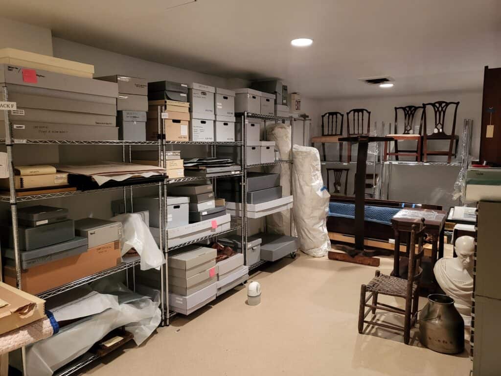 A storage room with shelves full of labeled boxes, wrapped items, folded chairs on an upper shelf, and miscellaneous furniture and objects.