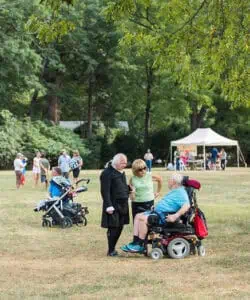 People gathered in a park; a man in historic clothing talks to an elderly man in a wheelchair, accompanied by a woman. Other individuals, including children, are in the background.
