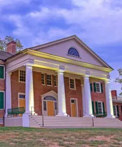 A brick building with white columns and green shutters, illuminated at dusk. The structure features a triangular pediment with a semicircular window. Steps lead up to the entrance with closed double doors.