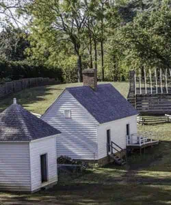 A small white house with a sloped roof and a chimney, adjacent to a round structure, set in a grassy area with trees and a wooden fence in the background.