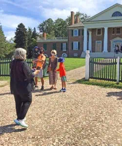 A group of people stands on a gravel path near a historic building with white columns and a porch, surrounded by a wooden fence and greenery.