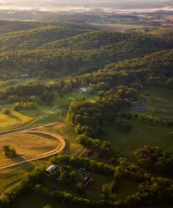 An aerial view of a rural landscape with dense greenery, scattered houses, and winding dirt roads during early morning light.