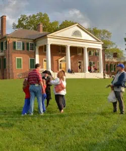 A group of people stand and chat on a grassy lawn in front of a large, historic brick building with white columns and green shutters.