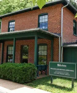 A two-story brick building with green trim, a porch, and a sign reading "William and Peyton Lewis Hall." Green bushes and a bench are in front.
