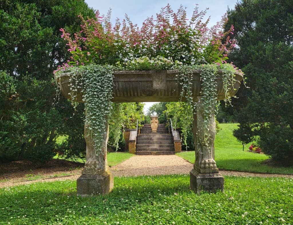 Stone planter with floral arrangements on top of two statues in a garden, with trees and stone steps leading to a pathway in the background.