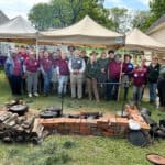 A group of people stands in front of a canopy tent near a cooking setup with firewood and pots. Buildings and trees are in the background.