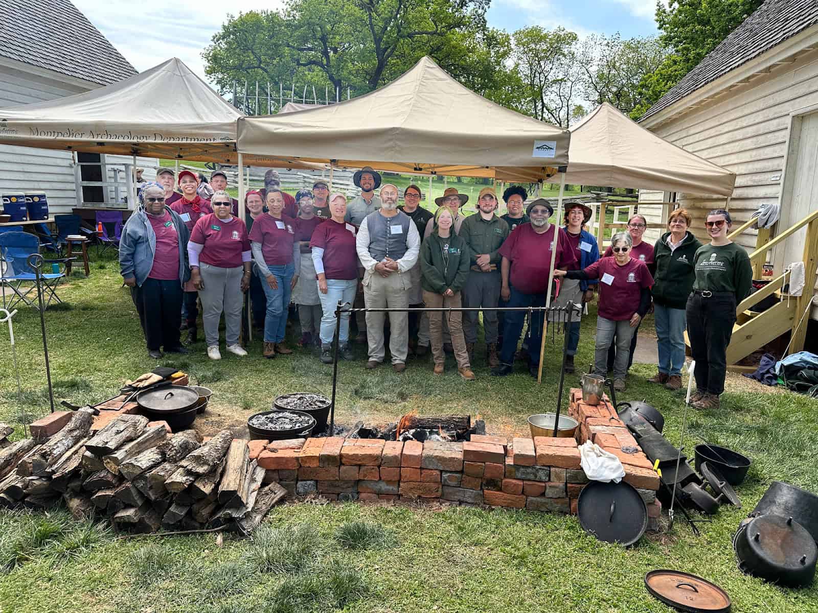 A group of people stands in front of a canopy tent near a cooking setup with firewood and pots. Buildings and trees are in the background.