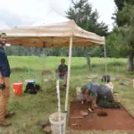 Three people at an outdoor archaeological dig site; one stands, one sits under a canopy, and one works on a square excavation pit in the ground.