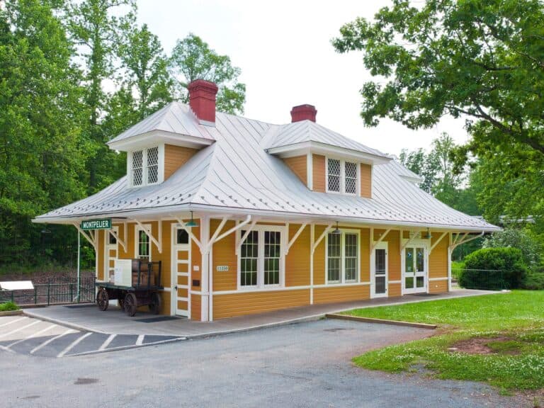 A historic yellow train station building with a metal roof, sign reading “Montpelier,” and an old luggage cart outside, surrounded by trees and greenery.