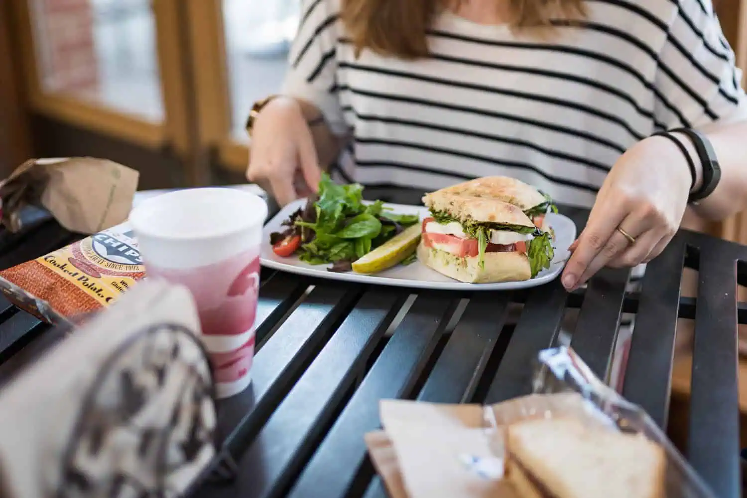 Closeup of a woman at a table with a sandwich, side salad and pickle on her plate with a bag of chips and a coffee on the side