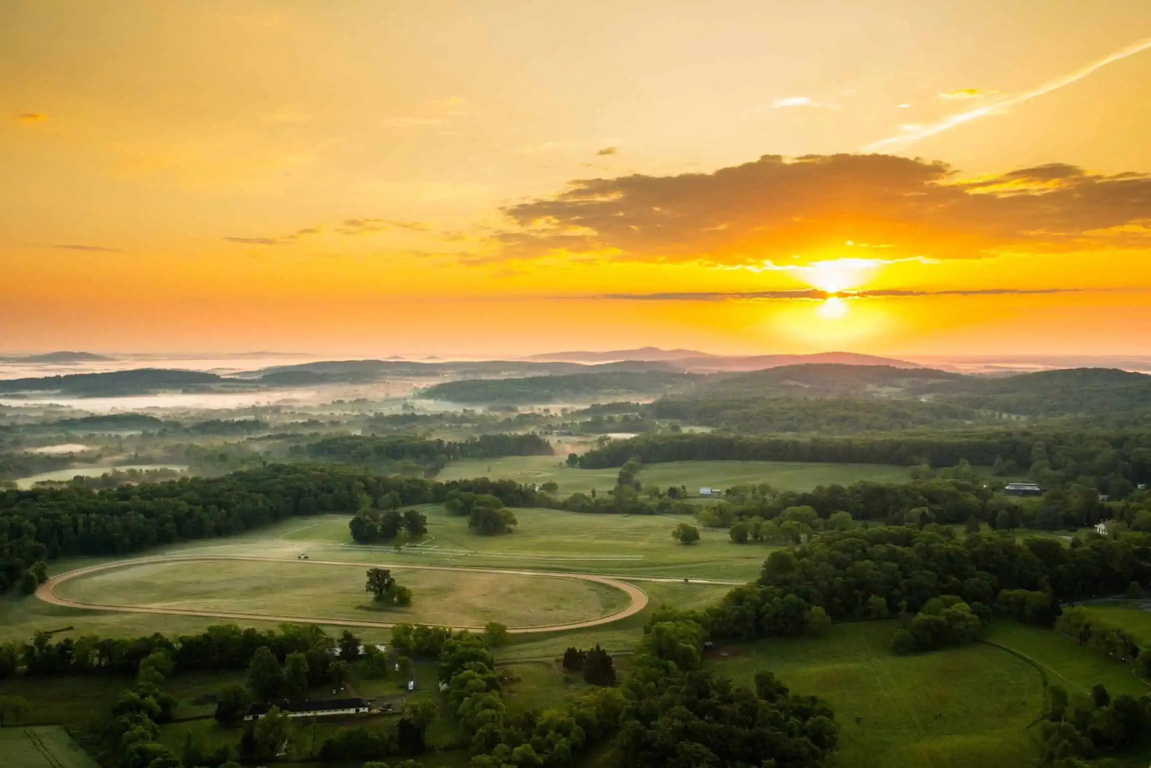 Aerial view of a lush green landscape in Orange County, VA, at sunrise, with a winding path and scattered trees under a glowing orange sky.