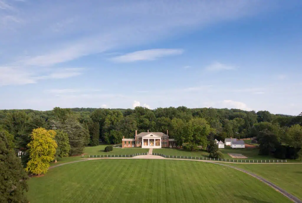 Large estate with a mansion surrounded by green lawns and trees, under a clear blue sky.