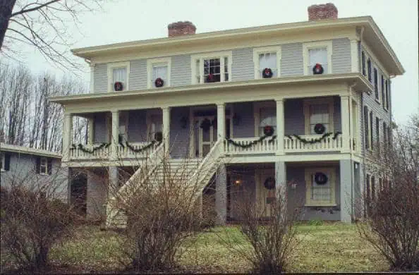 A two-story house with wreaths on windows and a porch decorated with garlands. Steps lead up to the entrance. Bare trees are in the background: The Exchange Hotel and Civil War Museum