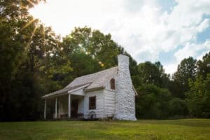 Small white cabin with a stone chimney, surrounded by lush greenery, under a bright sun and cloudy sky. Gilmore Cabin James Madison's Montpelier