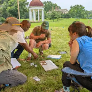 Four people kneel or sit on the grass, examining papers and artifacts outdoors near a domed pavilion and historic building in the background.