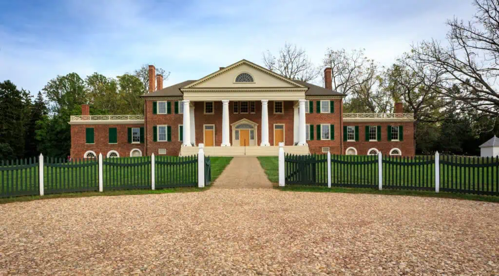 Front view of James Madison’s Montpelier, a red brick neoclassical mansion with white columns, green shutters, and a gravel walkway leading to the main entrance, surrounded by trees and fencing.