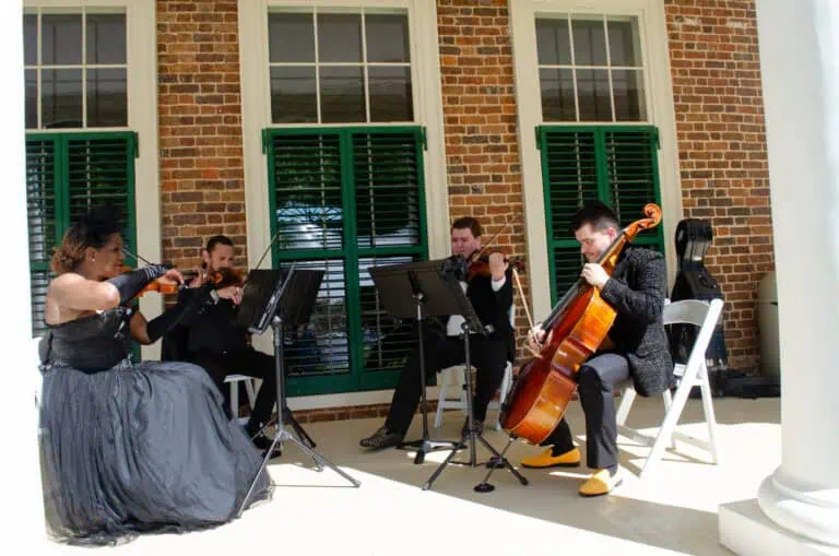 String quartet performing outdoors, featuring two violinists, one cellist, and one violist in formal attire, with brick wall and large windows in the background.