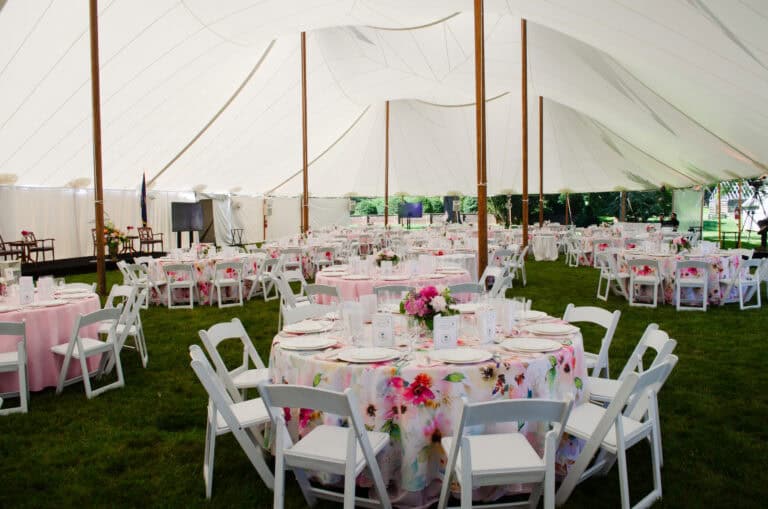 A wedding reception tent with round tables covered in floral tablecloths, set with white chairs and floral centerpieces on a grassy area.