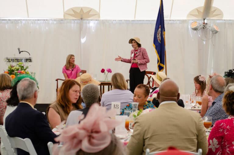 Two women speak on stage to a seated audience at an indoor event. The room is decorated with flowers and there's a flag in the background.