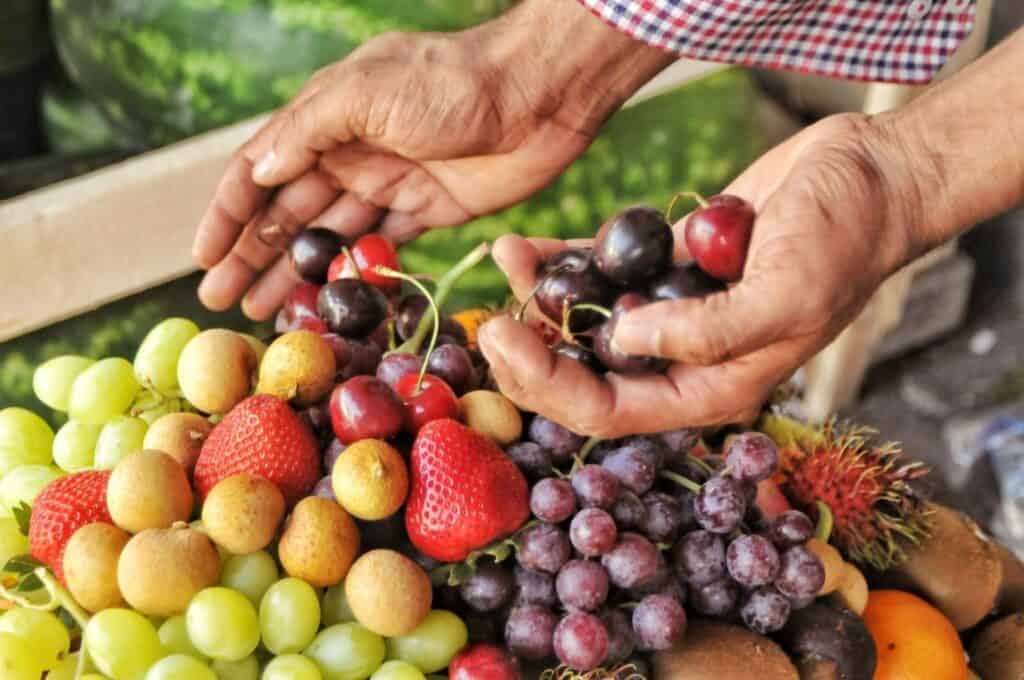 A person holding dark cherries above a colorful assortment of fresh fruits, including grapes, strawberries, longans, and kiwifruits.