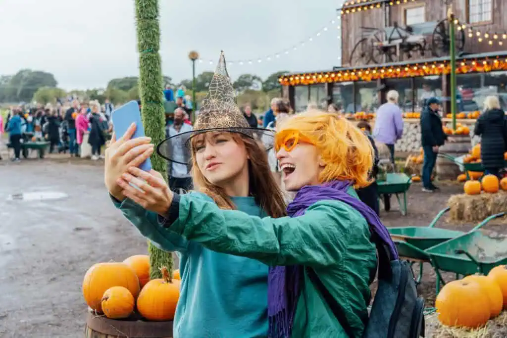 Two people take a selfie at a pumpkin patch festival, one in a witch hat and the other with orange hair and sunglasses. Pumpkins, lights, and crowds showcase the lively spirit of Festivals and Annual Events in Central Virginia.